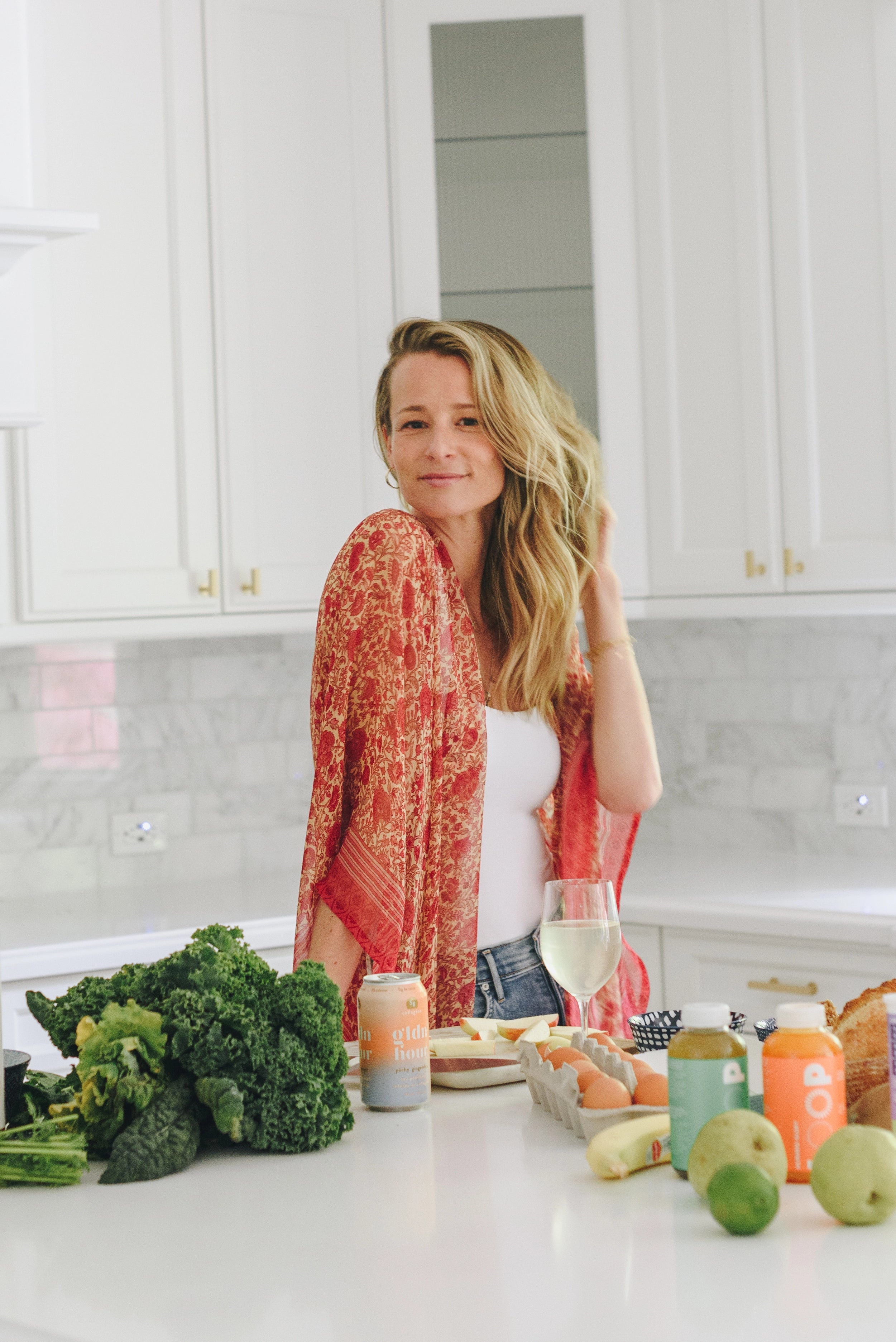 A woman in a red patterned kimono stands in a kitchen, smiling. She is surrounded by fresh produce, eggs, a loaf of bread, and canned beverages. A glass of white wine is on the counter. White cabinets and a marble backsplash are in the background.